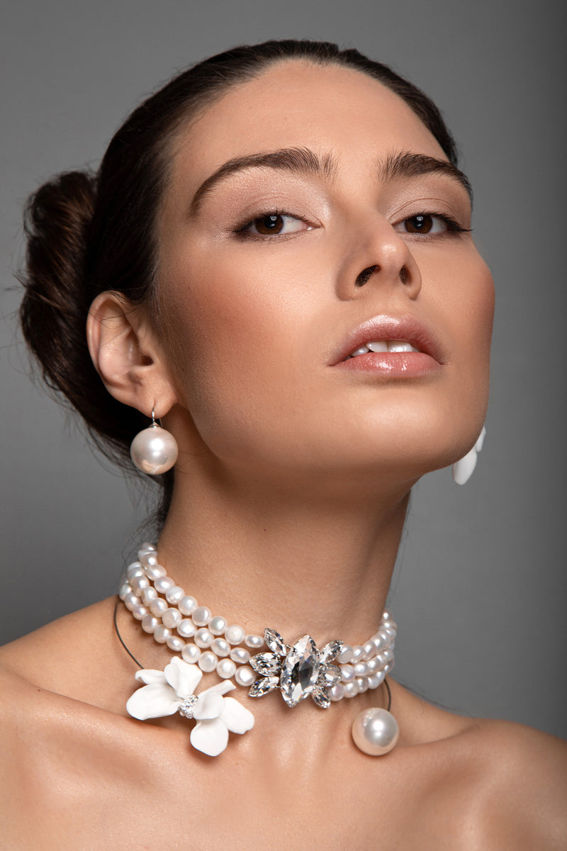 A close-up portrait of a woman wearing a multi-strand pearl Sildare jewlelry choker necklace with a white orchid flower and a sparkling crystal centerpiece, along with matching pearl drop earrings, against a solid gray background.
