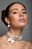 A close-up portrait of a woman wearing a multi-strand pearl Sildare jewlelry choker necklace with a white orchid flower and a sparkling crystal centerpiece, along with matching pearl drop earrings, against a solid gray background.