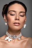 A close-up portrait of a woman wearing Sildare jewelry a pearl choker necklace with white flower charms and silver crystal detailing, along with matching pearl drop earrings, against a neutral gray background.