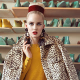 A model with blonde hair in a bun, wearing a leopard-print faux fur coat over a mustard yellow top, a red hat, and large black Sildare jewelry black handmade tassel earrings, standing in front of shelves of ceramic shoe boxes.