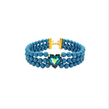 A double-strand beaded choker necklace in a medium blue color, featuring a central heart-shaped crystal charm with a blue-green gradient and gold settings, completed with a gold clasp.