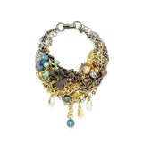 A multi-strand statement necklace featuring mixed metals including gold, silver, and bronze, adorned with faux pearls, blue beads, and dangling teardrop charms on a white background.